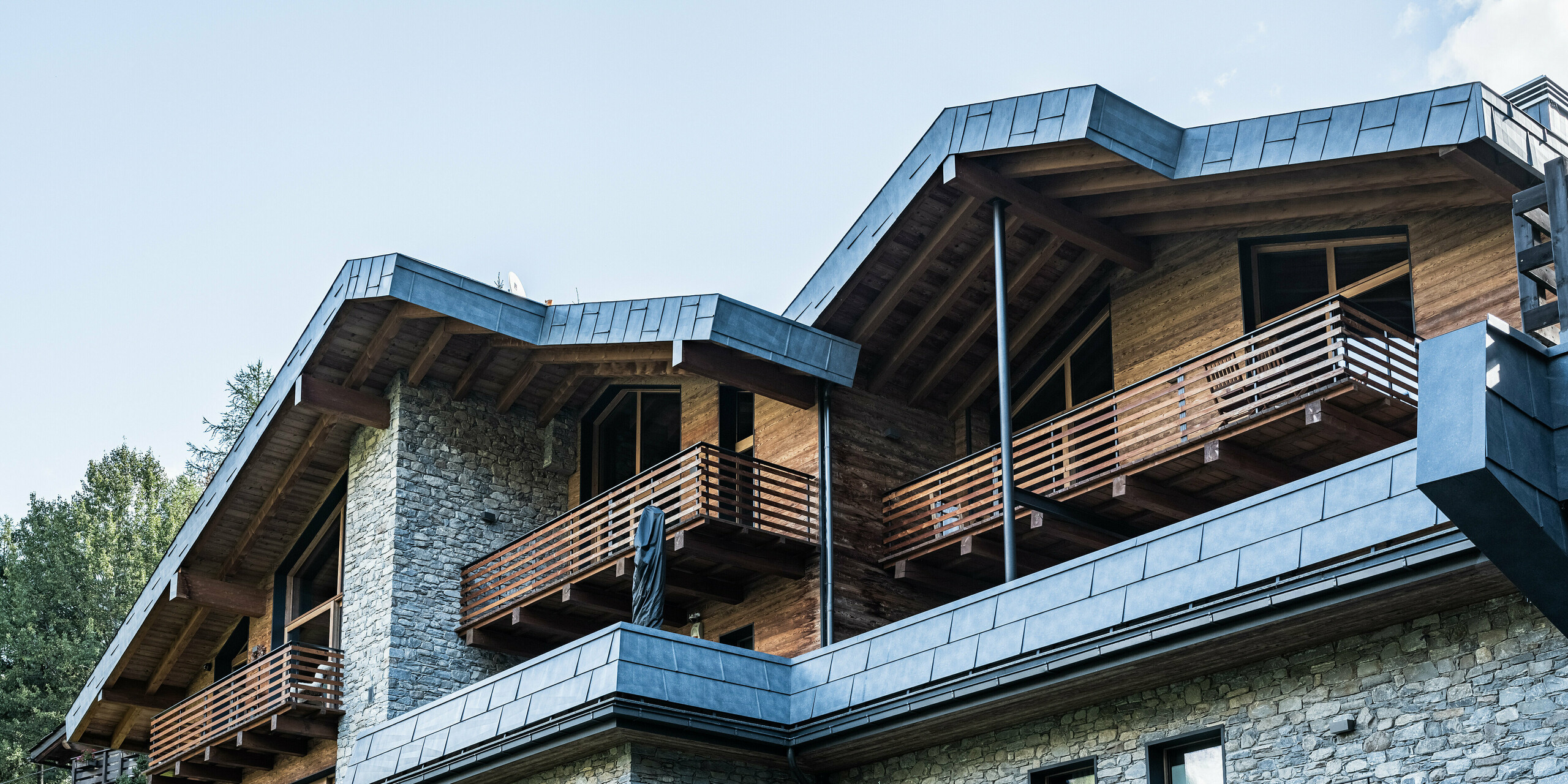 Detailaufnahme eines modernen Appartementhauses in Courmayeur mit Balkonen und einer Kombination aus Natursteinwänden und Holzverkleidung. Die markanten Dach- und Fassadenpaneele aus PREFA Aluminium in P.10 Steingrau unterstreichen die hochwertige Architektur. Eingerahmt wird das Gebäude von den umliegenden Pinien, die eine natürliche und ruhige Atmosphäre schaffen und das harmonische Zusammenspiel von modernen und traditionellen Bauelementen unterstreichen.