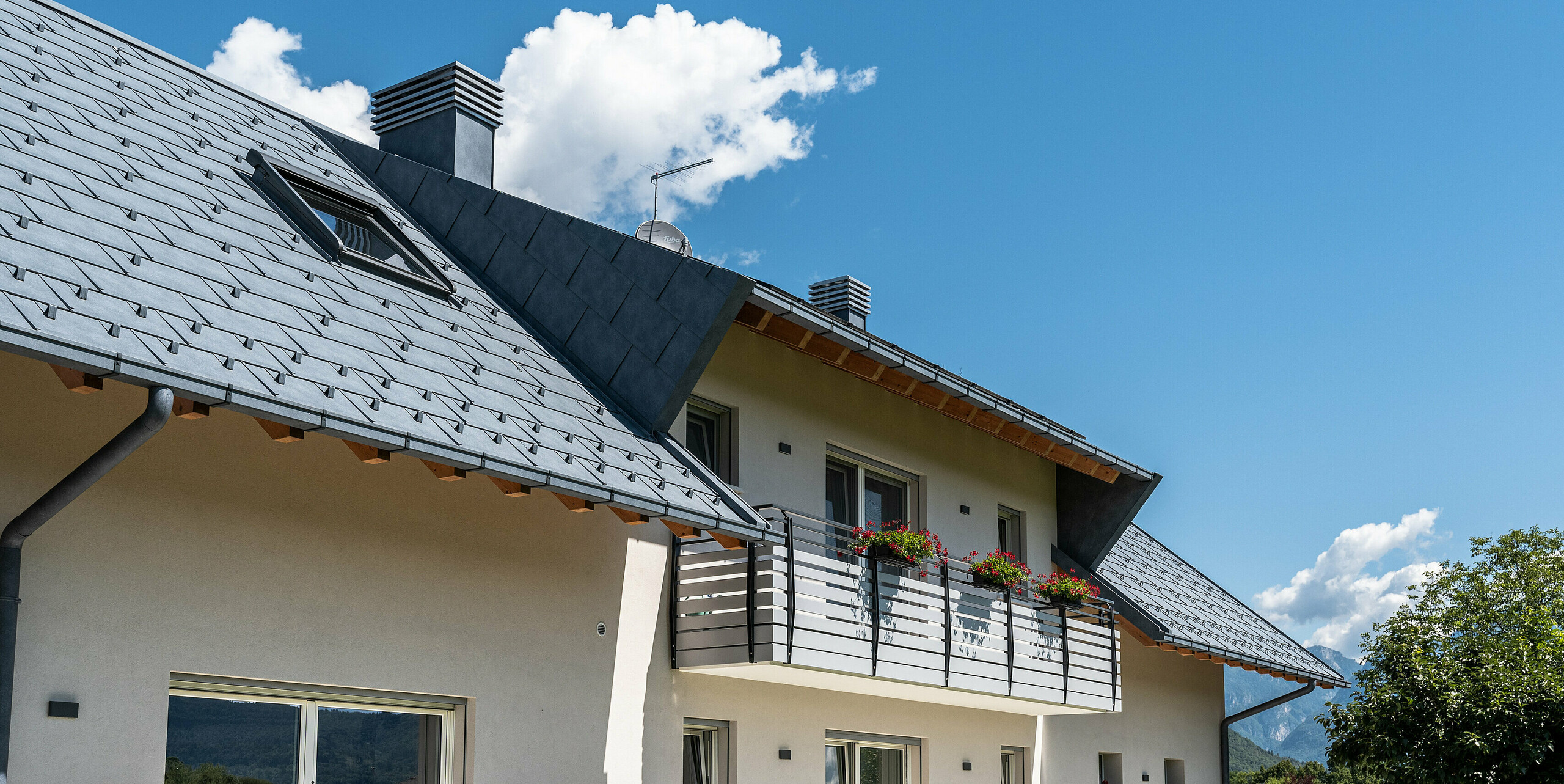 Ein Blick von unten auf das elegante Dach eines Hauses in Belluno, Italien. Die Dachfläche hat eine Schleppgaube und wurde mit PREFA R.16 Dachplatten in P.10 anthrazit eingedeckt. Die Dachentwässerung von PREFA, bestehend aus einer Kastenrinne und einem Ablaufrohr, ist farblich auf das robuste Aluminiumdach abgestimmt und schafft eine einheitliche und moderne Ästhetik bei optimalem Schutz.