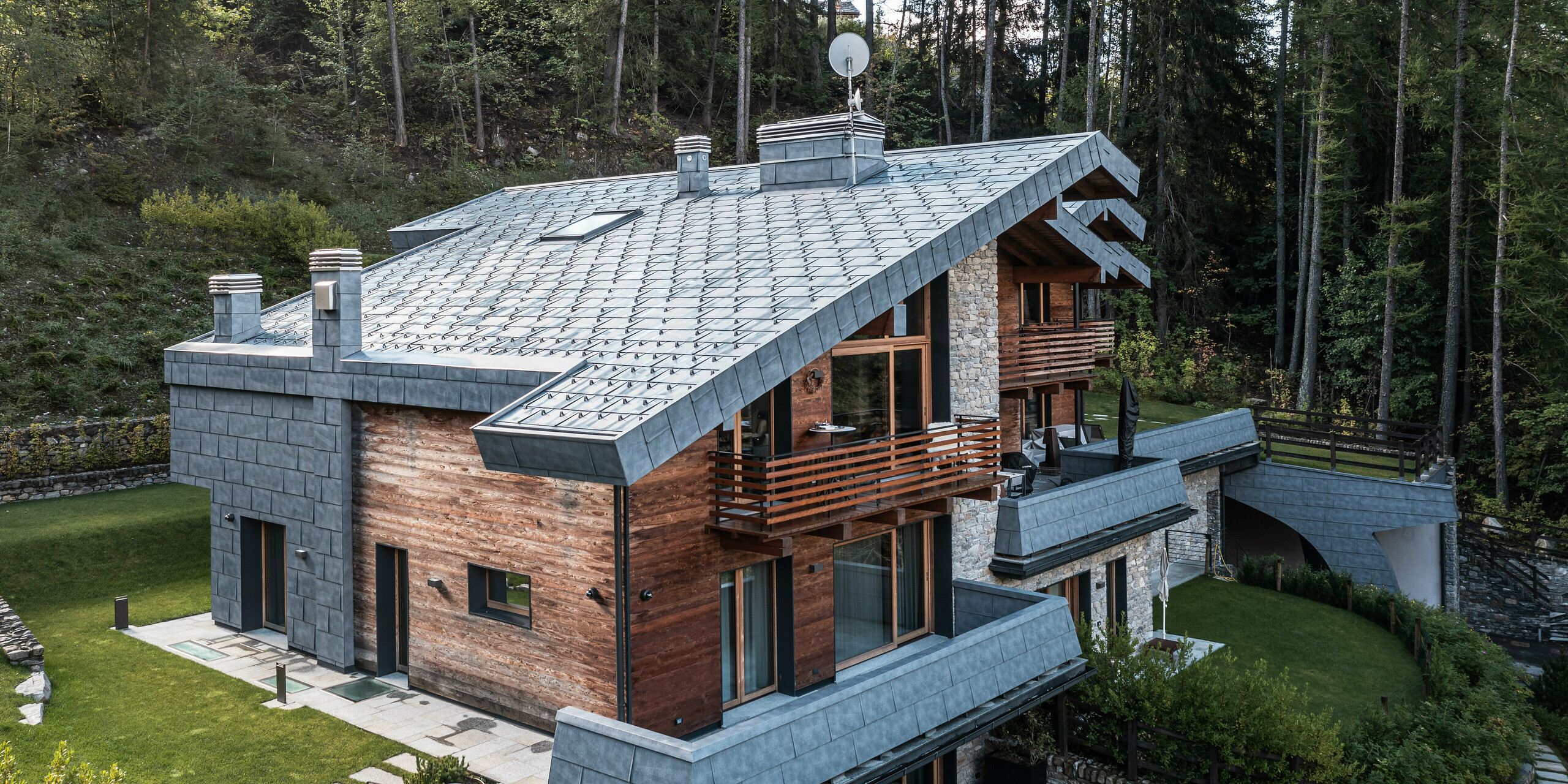 Die Vogelperspektive einer Luxusvilla in Courmayeur zeigt die beeindruckende Kombination aus Holz, Stein und PREFA FX.12 Aluminium-Dachpaneelen in Steingrau. Die robusten PREFA-Blechpaneele erstrecken sich vom Dach teilweise über die Fassade und bieten dem Gebäude einen starken Schutz vor Witterungseinflüssen. Die Architektur fügt sich nahtlos in die umgebende Waldlandschaft ein. Zu den Besonderheiten gehören mehrere Balkone und überdachte Terrassen, die einen entspannenden Blick auf die umliegende Natur bieten. Das Grundstück ist von einem liebevoll angelegten Garten umgeben, der die harmonische Einbindung in die alpine Landschaft unterstreicht.