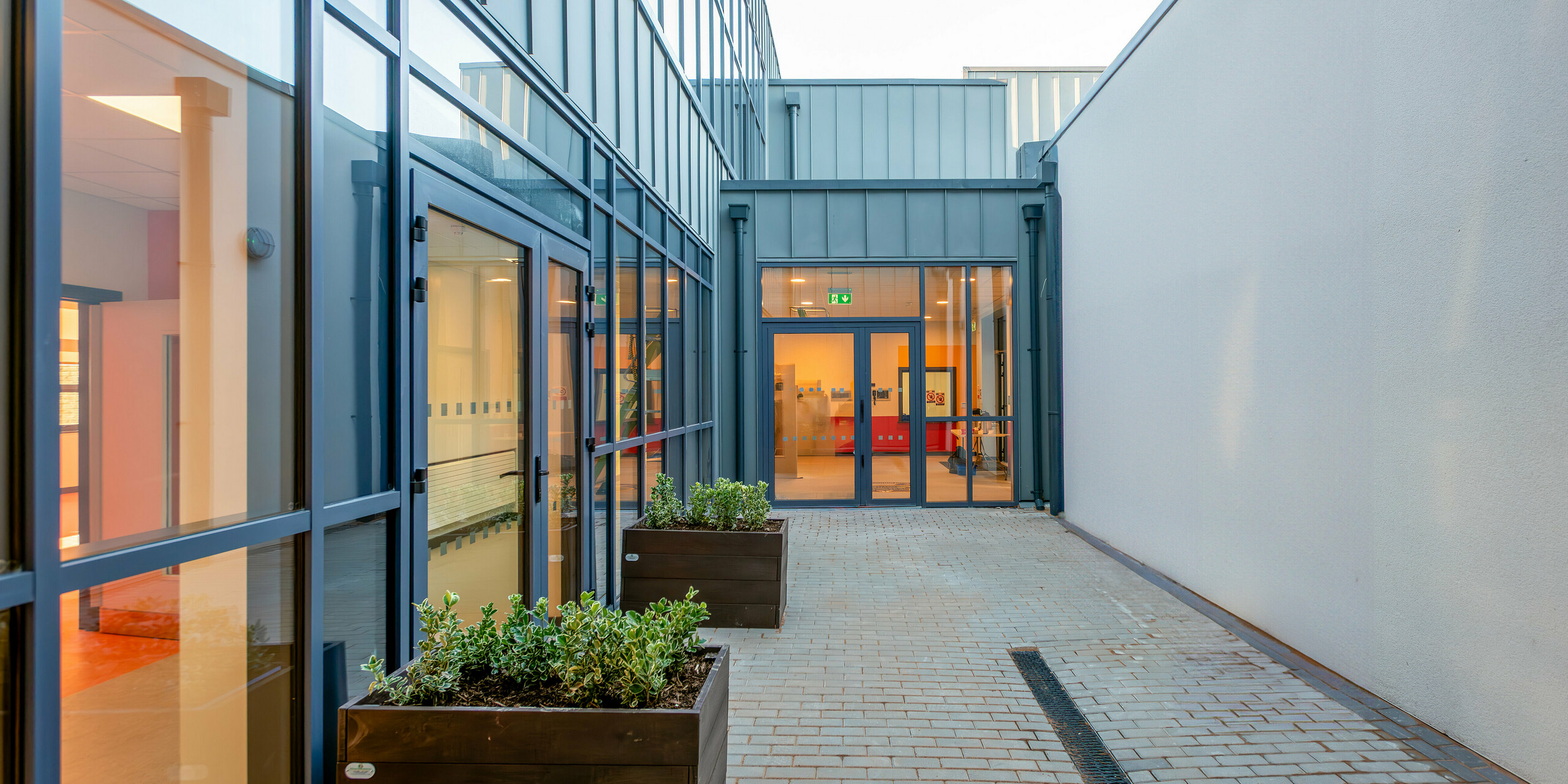 Moderner Durchgangsbereich der Scoil Mhuire Naofa in Carrigtwohill, Irland. Große Glasfronten und eine PREFALZ Aluminiumfassade in P.10 Hellgrau sorgen für eine lichtdurchflutete, zeitgemäße Architektur. Pflanzgefäße und helle Materialien schaffen eine einladende Atmosphäre.