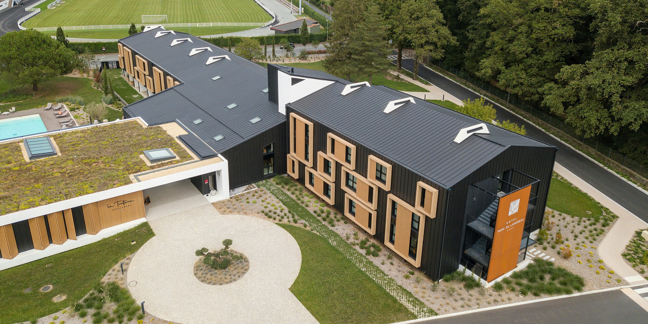 Luftaufnahme des Hôtel Parc du Landreau in Les Herbiers, Frankreich, mit einem schwarzgrauen PREFALZ Dach- und Fassadensystem. Die Kombination aus Holzrahmen um die Fenster und der hochwertigen Stehfalzverkleidung aus Aluminium von PREFA sorgt für ein elegantes Erscheinungsbild. Die begrünte Dachfläche über dem Eingangsbereich unterstreicht die nachhaltige Architektur des Hotels.