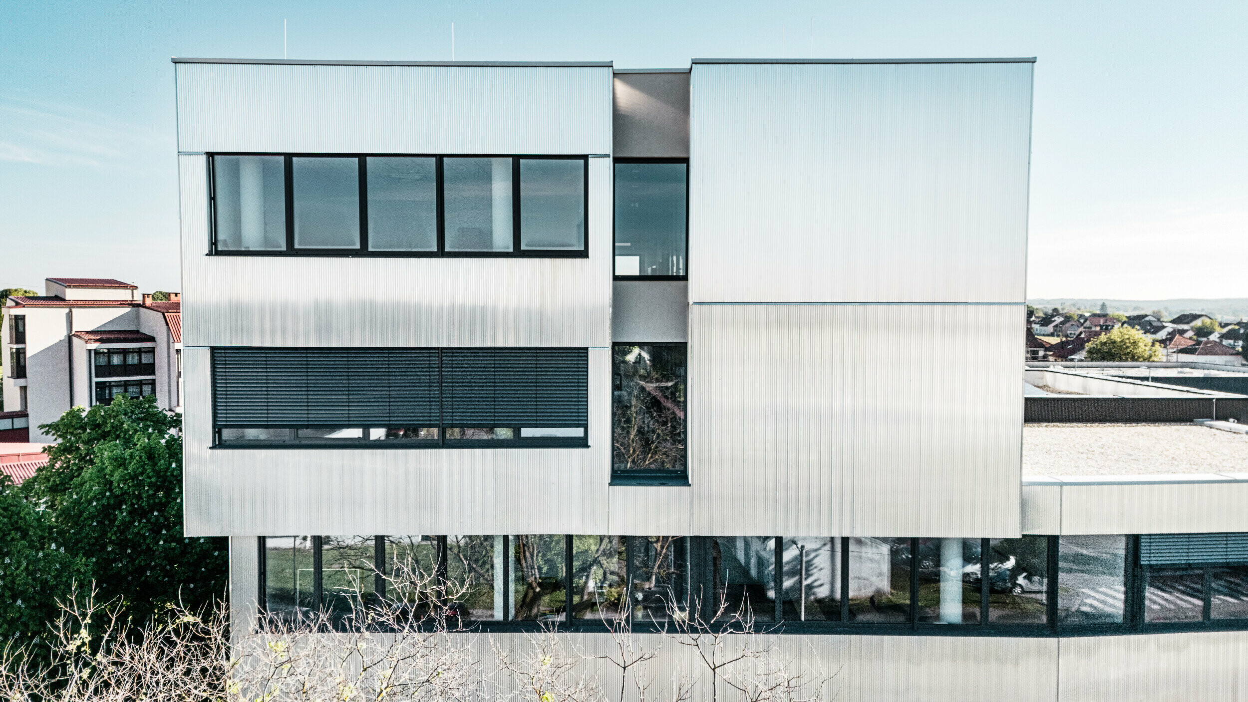 Eine moderne mehrstöckige Musikschule in Bjevolar mit einer PREFA Fassade im Zackenprofil. Die Fassade ist teilweise in Schwarz und aluminiumblank gehalten und reflektiert das Sonnenlicht, während große Glasfenster für einen modernen Look sorgen. Im Hintergrund sind Wohngebäude und Bäume sichtbar.