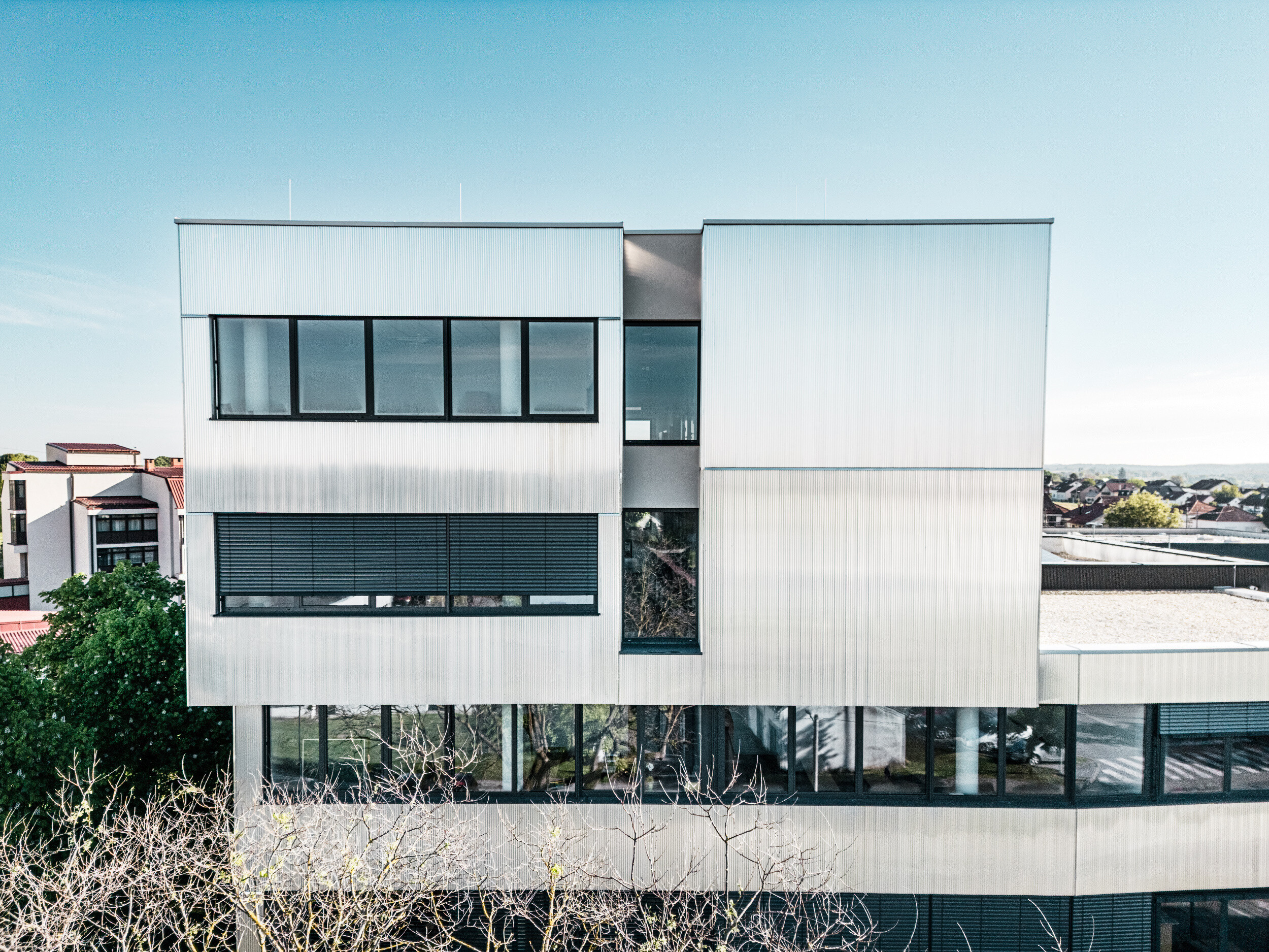 Eine moderne mehrstöckige Musikschule in Bjevolar mit einer PREFA Fassade im Zackenprofil. Die Fassade ist teilweise in Schwarz und aluminiumblank gehalten und reflektiert das Sonnenlicht, während große Glasfenster für einen modernen Look sorgen. Im Hintergrund sind Wohngebäude und Bäume sichtbar.