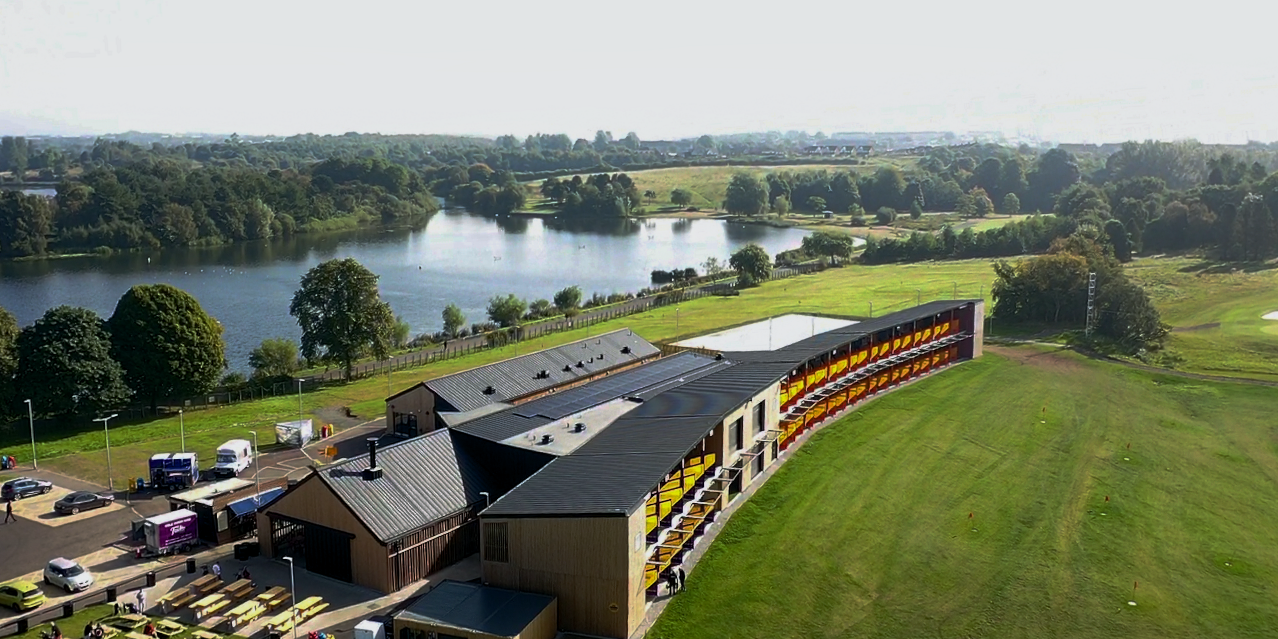 Drohnenaufnahme der Golfanlage 'Golf it!' in Glasgow, Schottland, mit einem PREFALZ Dachsystem in P.10 Schwarz. Die dunkle Dachgestaltung schafft einen eleganten Kontrast zu der Holzfassade des Gebäudekomplexes. Das Bauwerk fügt sich nahtlos in die umgebende Landschaft ein, mit einem klaren Blick auf den angrenzenden See und das üppige Grün des Golfplatzes. Die Dächer der einzelnen Bauteile mit ihrer einheitlichen, schwarzen Prefa Aluminiumverkleidung heben sich vor der natürlichen Kulisse ab und demonstrieren die harmonische Verbindung von moderner Architektur und landschaftlicher Schönheit.