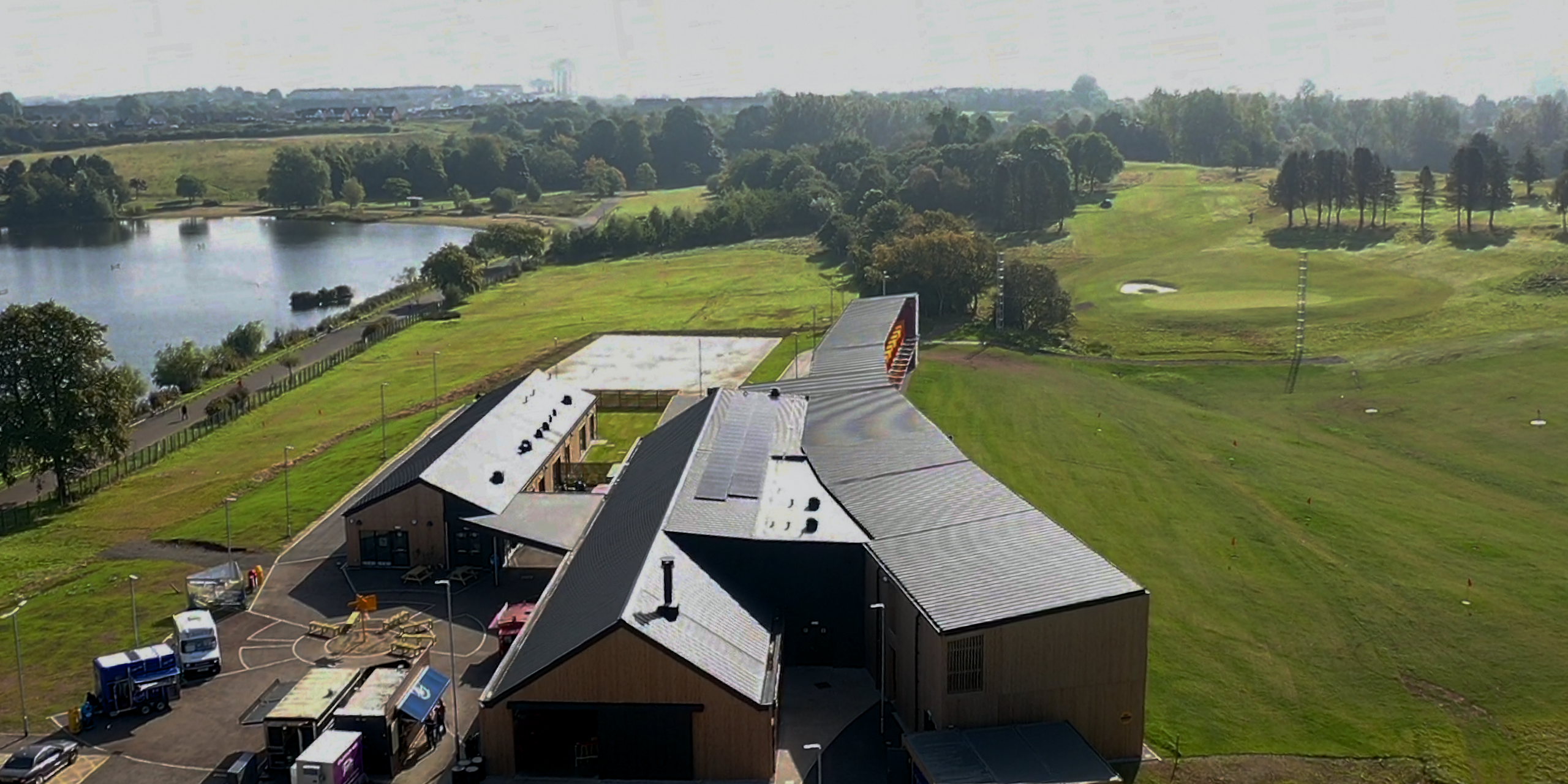 Luftaufnahme der 'Golf it!' Golfanlage in Glasgow, die inmitten einer malerischen Landschaft mit einem See und einem weitläufigen Golfplatz liegt. Die Gebäude sind mit PREFALZ in P.10 Schwarz gedeckt. Diese Ansicht demonstriert die Anpassungsfähigkeit und ästhetische Qualität von PREFA Aluminiumprodukten in einer natürlichen Umgebung.