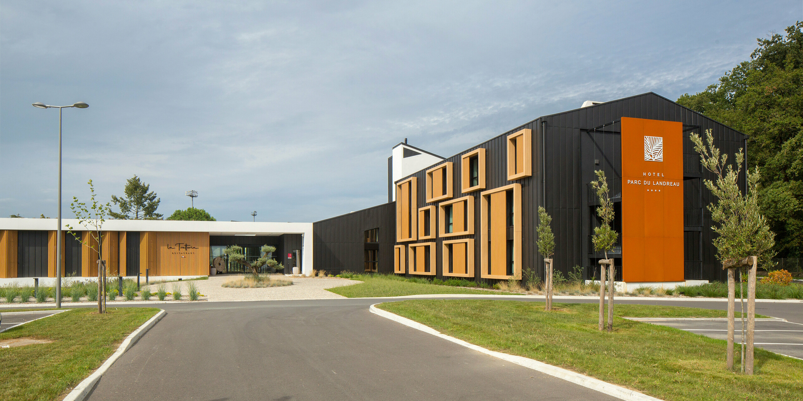 Zugang vom Parkplatz zum Hôtel Parc du Landreau in Les Herbiers, Frankreich. Das moderne Gebäude ist mit dem PREFALZ Dach- und Fassadensystem in Schwarzgrau verkleidet. Die moderne Architektur wird durch markante Holzrahmen um die Fenster ergänzt, die sich harmonisch in die robuste Aluminiumverkleidung einfügen. Der großzügige Vorplatz, die gepflegten Grünflächen und das zeitlose Design unterstreichen die einladende Atmosphäre des Hotels.