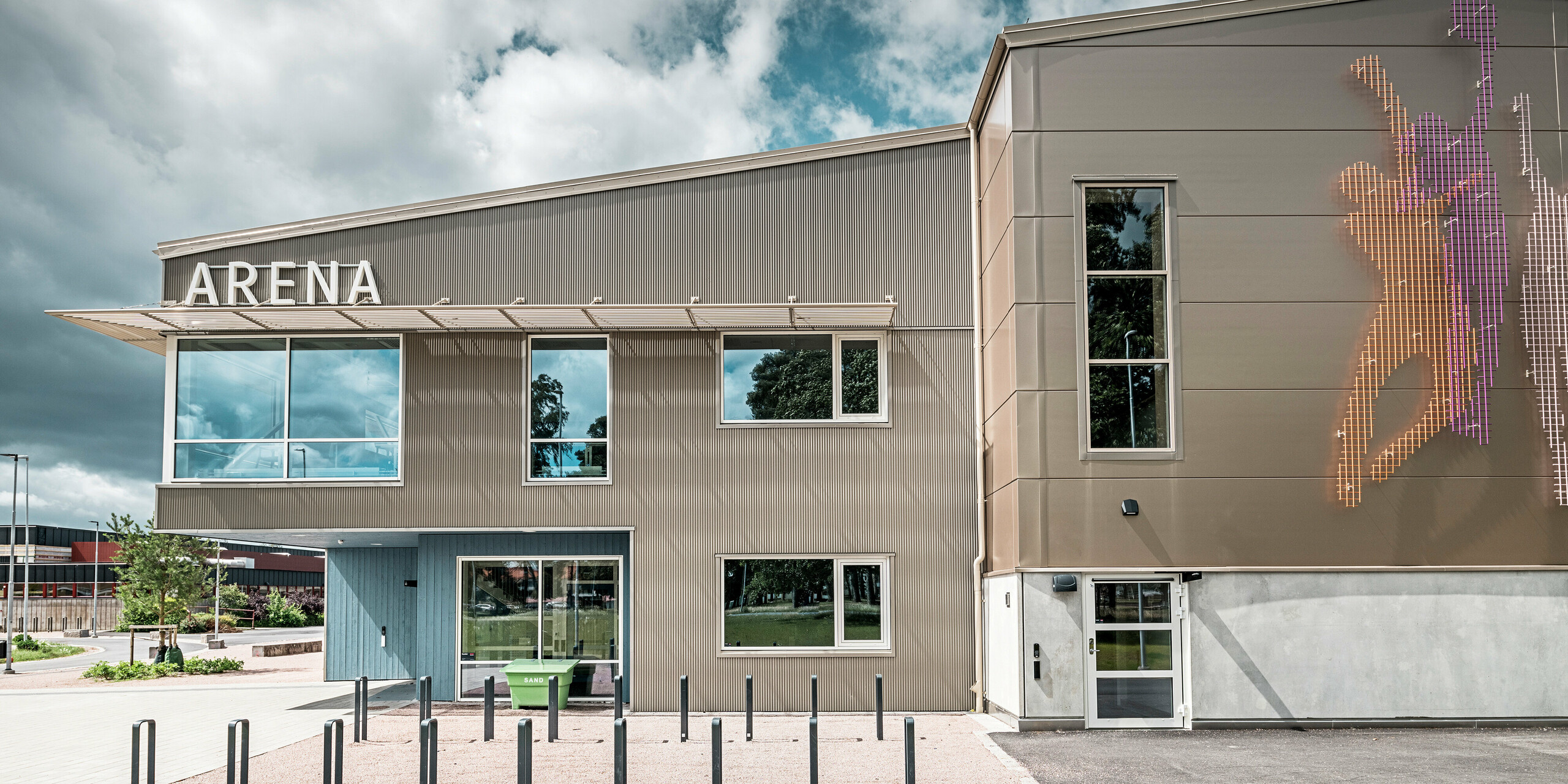 Seitenansicht des Eingangsbereichs zur Park Arena in Värnamo, Schweden, mit moderner PREFA Fassade aus dem Zackenprofil in Bronze. Die Aluminiumfassade ist langlebig und sorgt für markante Licht- und Schatteneffekte. Rechts im Bild befindet sich ein künstlerisch gestaltetes Wandbild, das sportliche Figuren darstellt und die dynamische Atmosphäre der Sporthalle unterstreicht. Die großen Fensterflächen lassen viel Licht in das Gebäude und betonen das zeitgemäße Design.