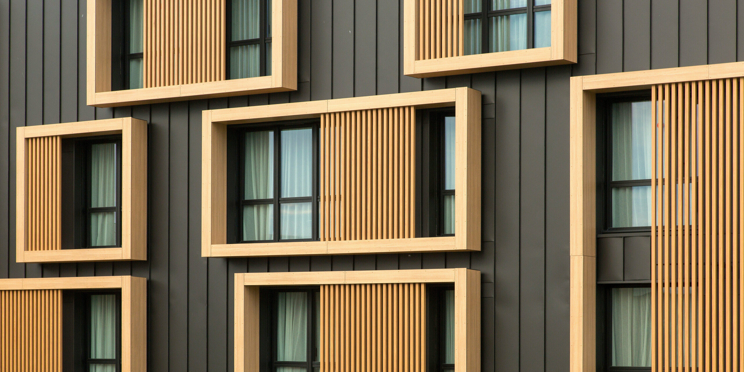 Nahaufnahme der Fassade des Hôtel Parc du Landreau in Les Herbiers, Frankreich, mit einer Kombination aus schwarzgrauem PREFALZ und Holzrahmen um die Fenster. Die vertikale Stehfalzverkleidung aus Aluminium wird durch die hölzernen Fensterrahmen ergänzt, die der modernen Architektur einen warmen und natürlichen Akzent verleihen.