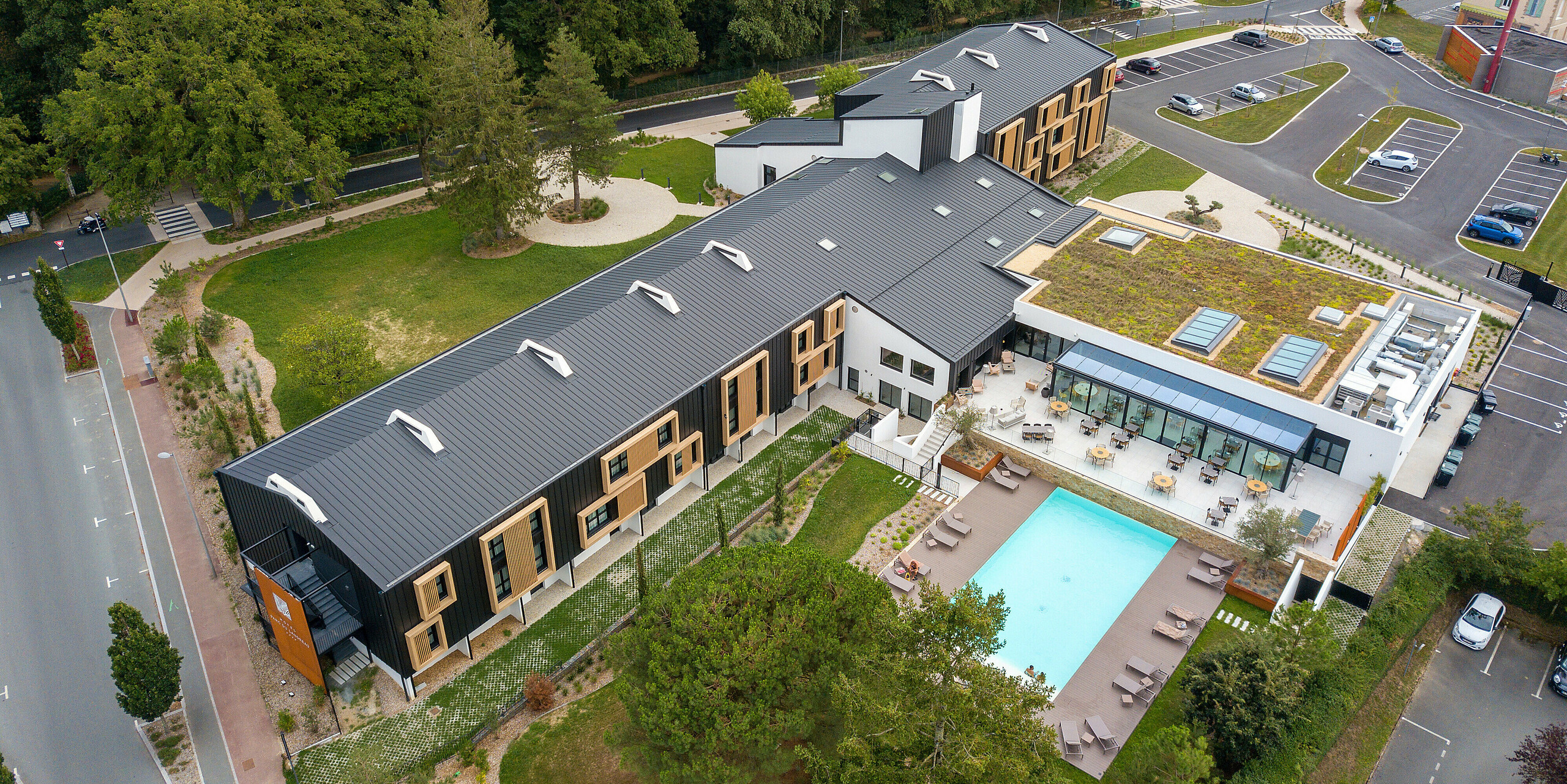 Luftaufnahme der Rückseite des Hôtel Parc du Landreau in Les Herbiers, Frankreich, mit dem PREFALZ Dach- und Fassadensystem in Schwarzgrau. Naturbelassene Holzelemente um die Fenster unterstreichen das moderne Gestaltungskonzept. Ein Gebäudeteil wurde mit einem begrünten Dach ausgestattet, das die langlebige Aluminium-Stehfalzdeckung optisch ergänzt. Der großzügige Außenbereich mit Pool und Terrasse bietet den Gästen eine einladende Atmosphäre.