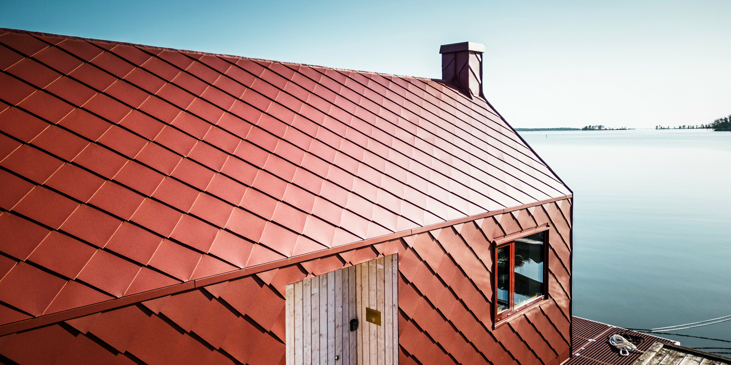 Architektonische Detailaufnahme des schwimmenden Stationshauses der Seenotretter am Hjälmaren See, mit einer spektakulären PREFA Aluminiumhülle aus Dach- und Wandrauten 29×29 in P.10 Oxydrot. Die präzise angeordneten Rauten am Dach und der Fassade spiegeln die Beständigkeit und Widerstandsfähigkeit von PREFA wider und verleihen dem Gebäude ein modernes und sympathisches Erscheinungsbild. Das schwimmende Stationshaus muss etwa die Hälfte des Jahres herrschendem Eis trotzen und ist optimiert für die hohe Beanspruchung durch Wind und Wellen.