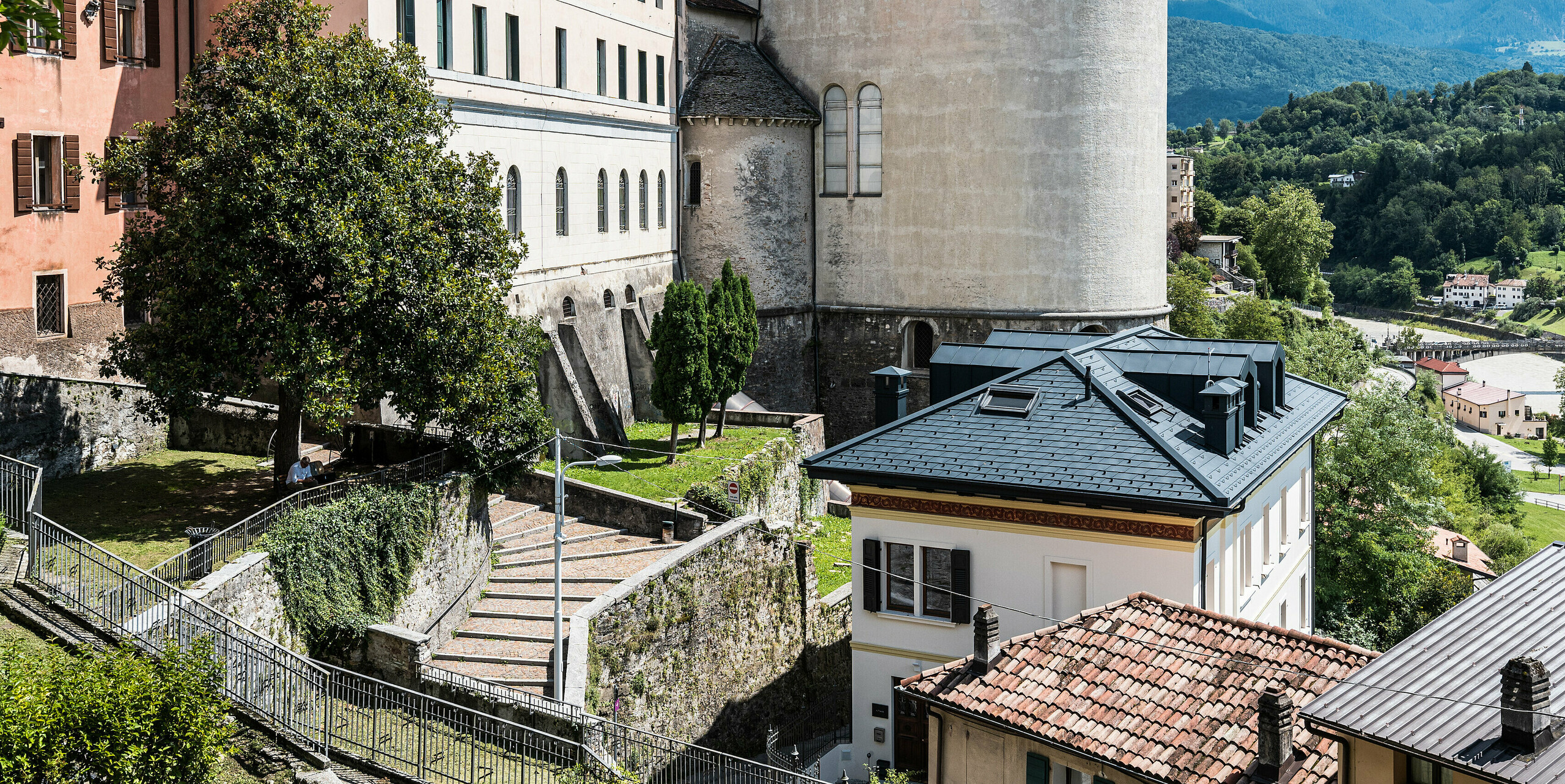 Idyllische Ansicht des historischen Zentrums von Belluno, Italien, mit einem sorgfältig restaurierten Gebäude, dessen Dach mit PREFA Aluminiumdachschindeln in Anthrazit ausgestattet ist, das harmonisch mit der klassischen Architektur und der umgebenden grünen Landschaft verschmilzt. Die Renovierung achtet auf die Wahrung der historischen Substanz, während sie moderne Elemente integriert, um das Erbe für zukünftige Generationen zu erhalten.