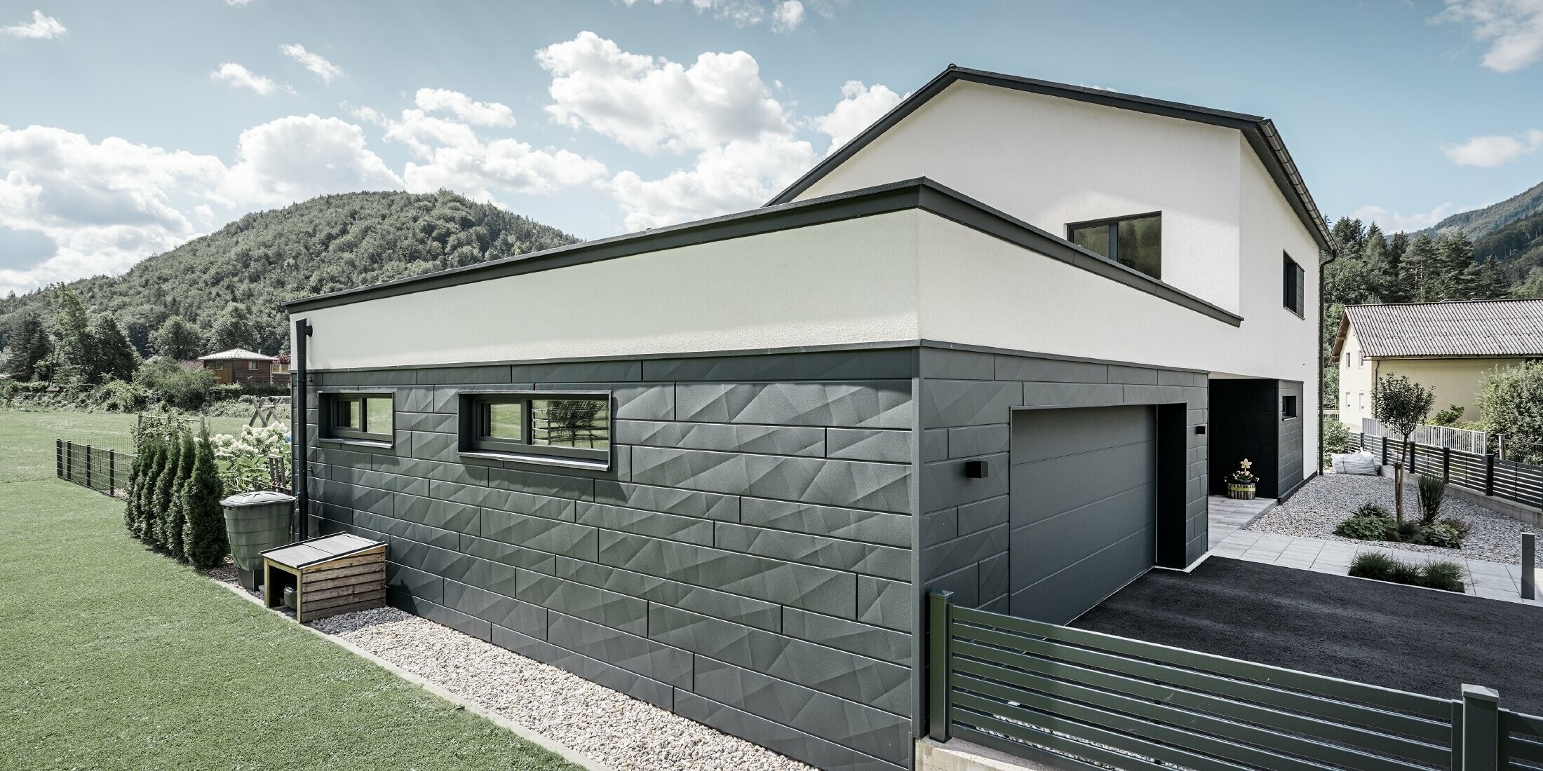 Das Foto zeigt die Seitenansicht eines modern gestalteten Einfamilienhauses mit einer Garage. Die Fassade ist überwiegend mit PREFA Siding.X in P.10 Anthrazit verkleidet, was der Immobilie ein zeitgenössisches und elegantes Erscheinungsbild verleiht. Die anthrazitfarbenen Fassadenelemente aus Aluminium harmonieren hervorragend mit den weißen Putzbereichen und der grauen Garagentür, wodurch ein stimmiges Farbkonzept entsteht. Der gepflegte Rasen und die Kieswege runden das ästhetische Bild des Grundstücks ab und betonen die ruhige, natürliche Umgebung mit den bewaldeten Hügeln im Hintergrund.