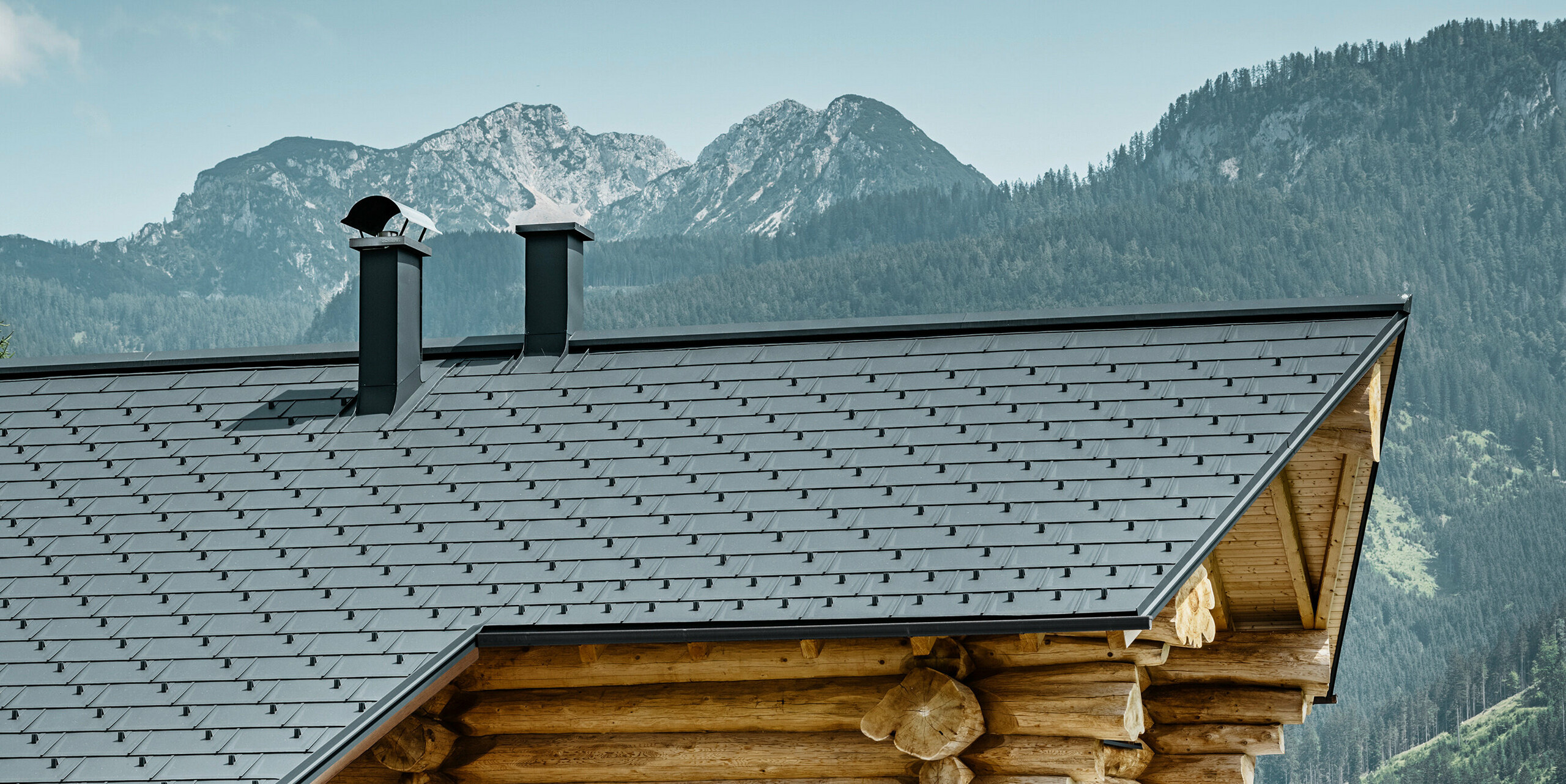 Eindrucksvolle Ansicht eines traditionellen Blockhauses in Gosau, Österreich, vor einem beeindruckenden Bergpanorama. Das Haus verfügt über ein PREFA Aluminiumdach bestehend aus Dachplatten R.16 in P.10 Anthrazit. Das anthrazitfarbene Dach bildet einen modernen Akzent zur rustikalen Holzbauweise. Die hochwertigen Dachplatten fügen sich nahtlos in die alpine Landschaft ein, während die Natürlichkeit des Holzhauses den ländlichen Lebensstil unterstreicht. Die Kombination aus natürlichen Materialien und dem ästhetischen Dachdesign verleiht diesem Anwesen einen zeitlosen Charme.