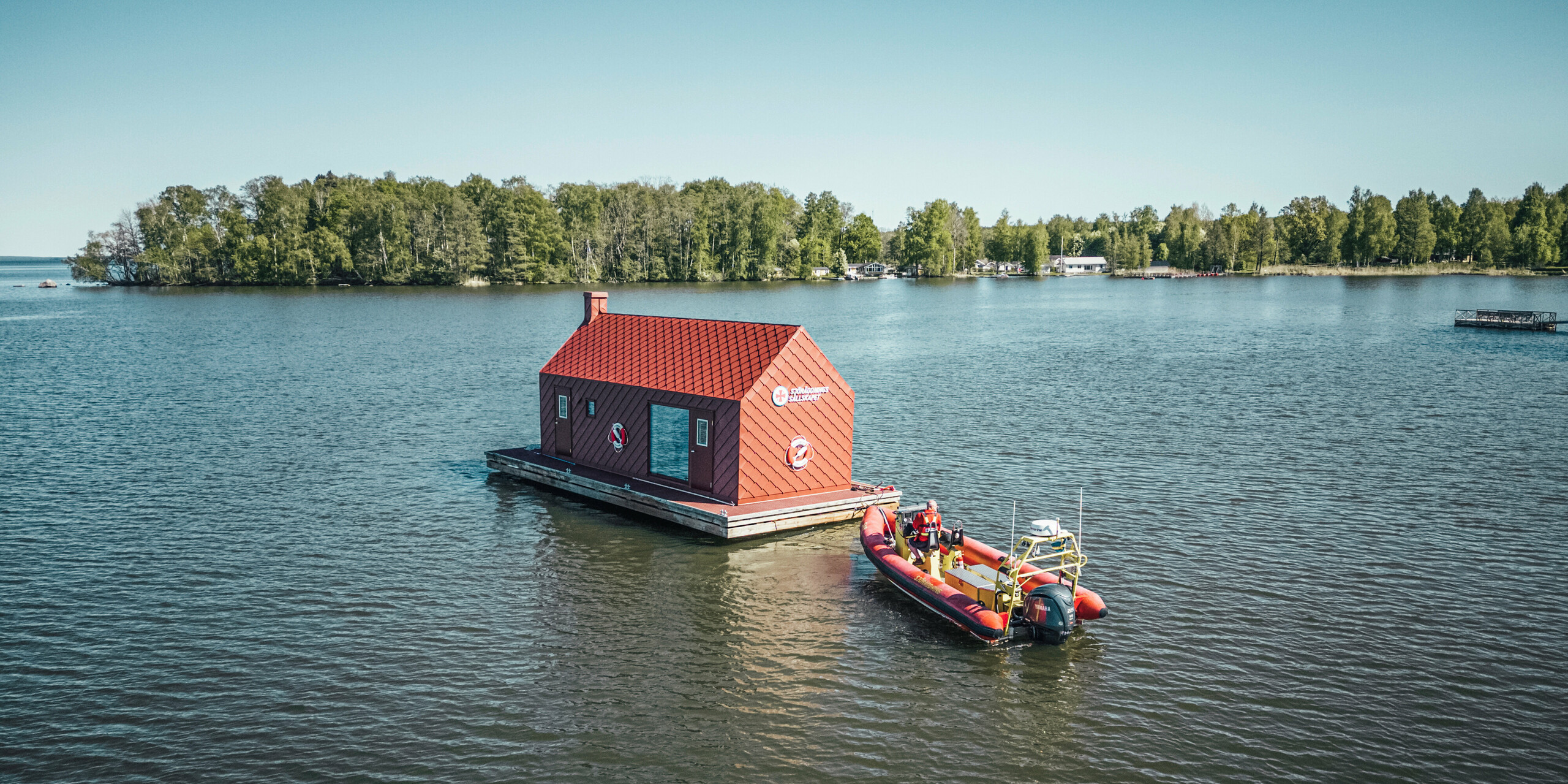 Weitwinkelaufnahme des Seenotrettungsstationshauses auf dem Hjälmaren See, schwimmend auf einer ruhigen Wasserfläche, mit einem Rettungsboot im Vordergrund. Das Gebäude ist mit einer oxydroten PREFA Aluminiumverkleidung aus Dach- und Wandrauten 29×29 bedeckt, was ihm eine lebhafte Ausstrahlung und robuste Haltbarkeit verleiht. Es dient als unverzichtbarer Stützpunkt für die Seenotretter, die auf diesem großen schwedischen See für Sicherheit sorgen.