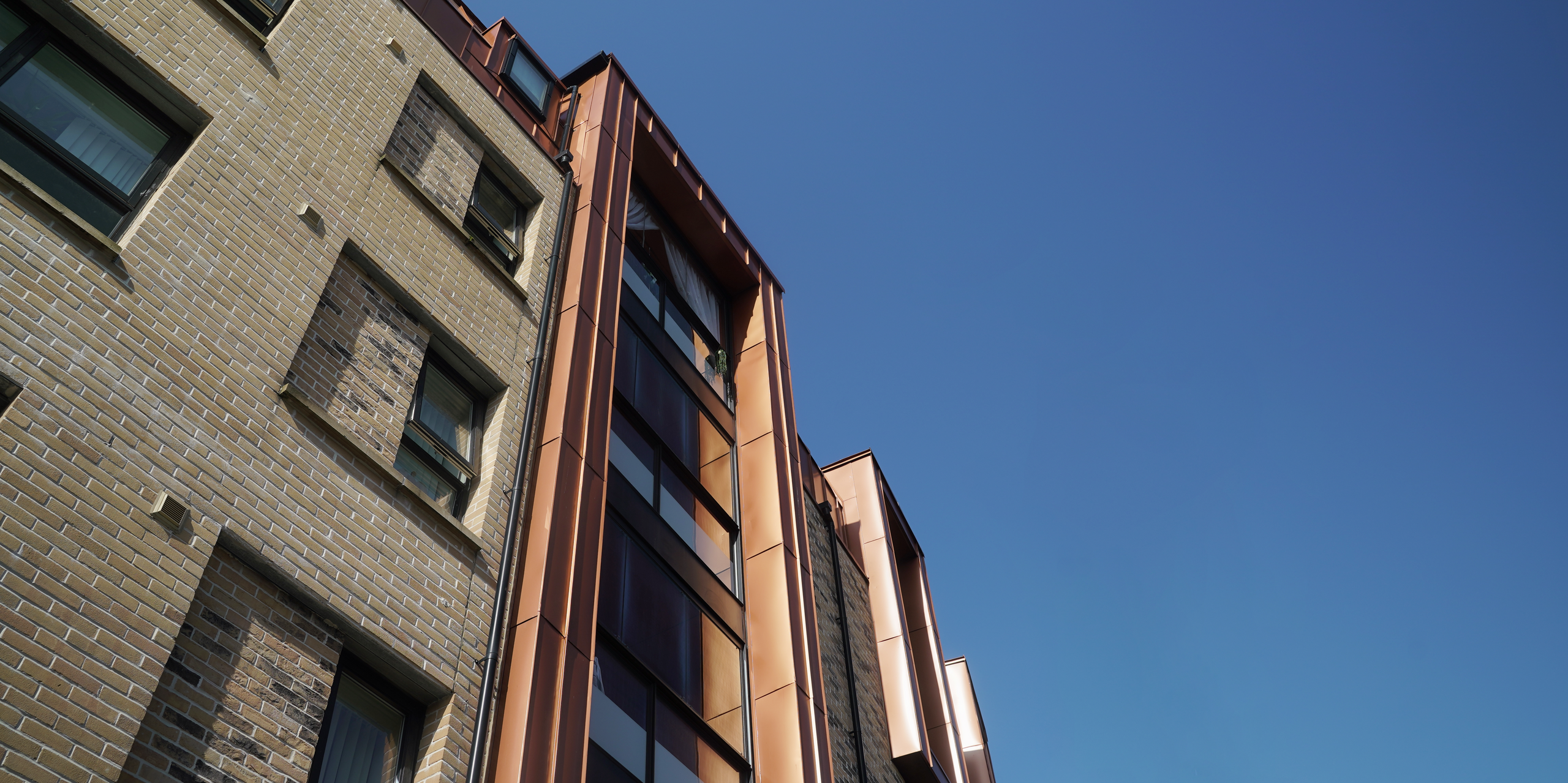 Perspektivische Ansicht der Fassade einer modernen Wohnhausanlage in der Nethan Street in Glasgow, verkleidet mit FALZONAL in Neukupfer. Die metallischen Fassadenelemente fangen das Licht ein und reflektierten es, was die Gebäudeform betont und einen eindrucksvollen visuellen Kontrast zur traditionellen Backsteinarchitektur bietet. Das PREFA Aluminiumprodukt sorgt nicht nur für eine ansprechende Ästhetik, sondern verspricht auch Langlebigkeit und geringen Wartungsaufwand.