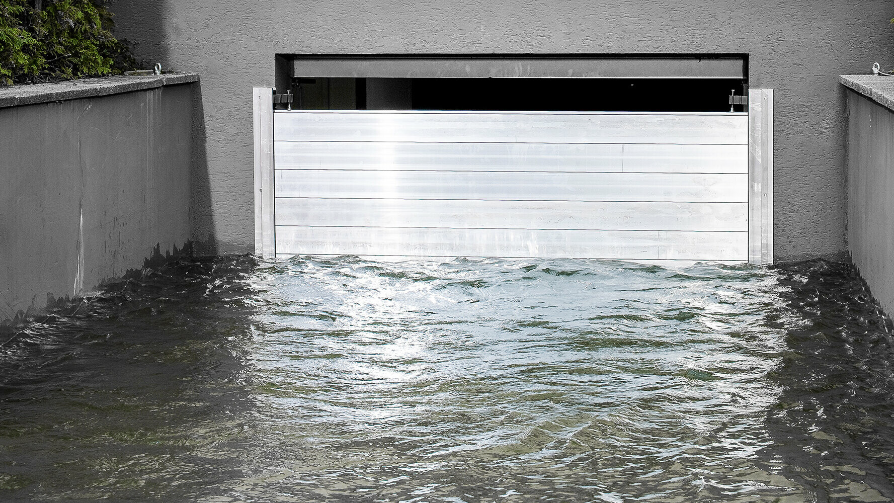 PREFA zaštita od poplava na djelu: Aluminijska barijera učinkovito štiti od poplava. Sjajni metalni zid prkosi uzburkanoj vodi i štiti garažu iza sebe. Ovo HWS rješenje kombinira funkcionalnost s modernim, estetskim dizajnom i demonstrira održivu zaštitu objekata i krajolika snagom PREFA-e.