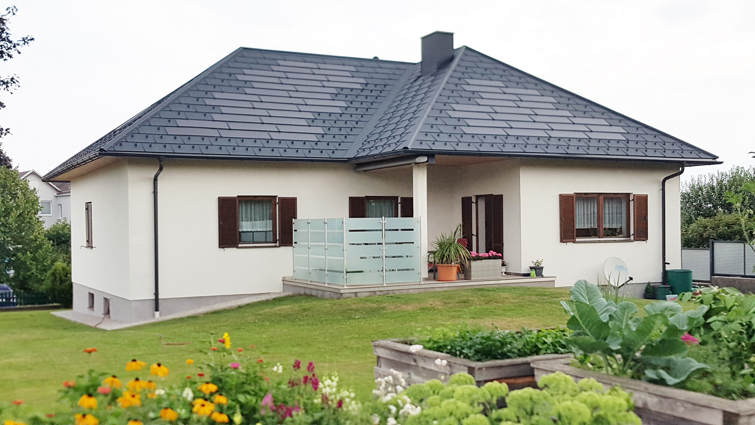 Ein klassisches Einfamilienhaus in Weitra, Niederösterreich, das mit den innovativen PREFA Solardachplatten und Dachplatten R.16 saniert wurde. Das Dach besticht durch seine moderne und elegante Optik, kombiniert mit fortschrittlicher Solartechnologie zur umweltfreundlichen Energiegewinnung. Das Haus hat eine beige Putzfassade und braune Fensterläden, die einen schönen Kontrast zu der Dachbedeckung bilden. Im Vordergrund ist ein gepflegter Garten mit bunten Blumen und Gemüsebeeten zu sehen, der die idyllische Atmosphäre des Grundstücks unterstreicht. Die hochwertige PREFA-Bedachung sorgt für Langlebigkeit, Witterungsbeständigkeit und Energieeffizienz.