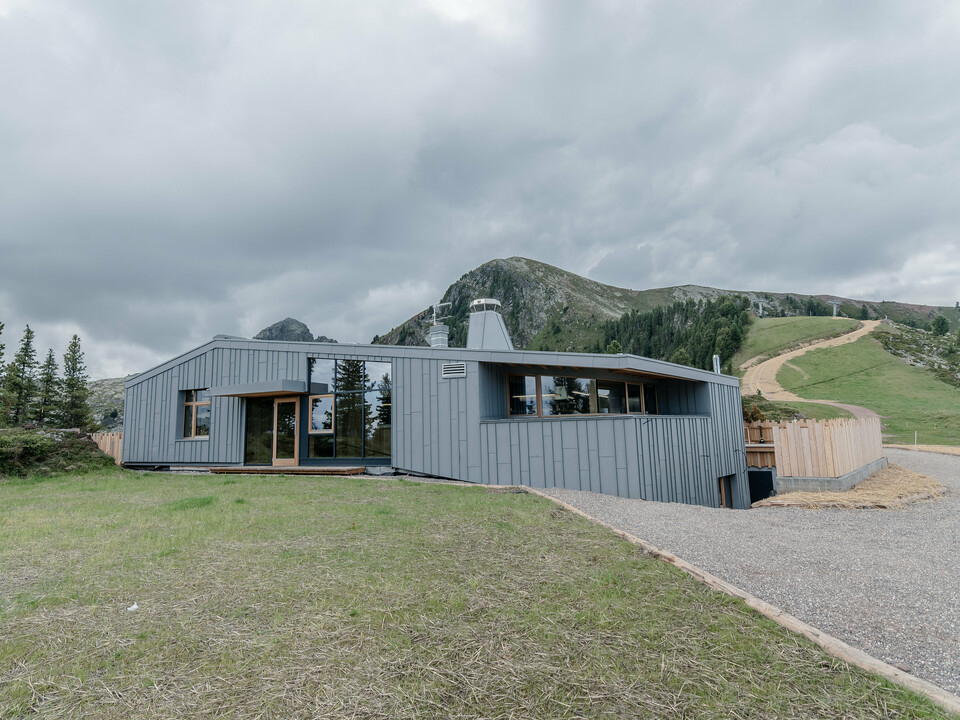 Das Ristorante Busabelle, eingebettet in eine idyllische Berglandschaft, ist von einer Winkelstehfalz Spiegeldeckung aus hellgrauem PREFALZ ummantelt, die die moderne Architektur hervorhebt und eine harmonische Verbindung zur natürlichen Umgebung schafft. Der Eingangsbereich ist mit großen Glasflächen versehen. Im Hintergrund sind Masten für eine Sesselliftanlage zu erkennen. 