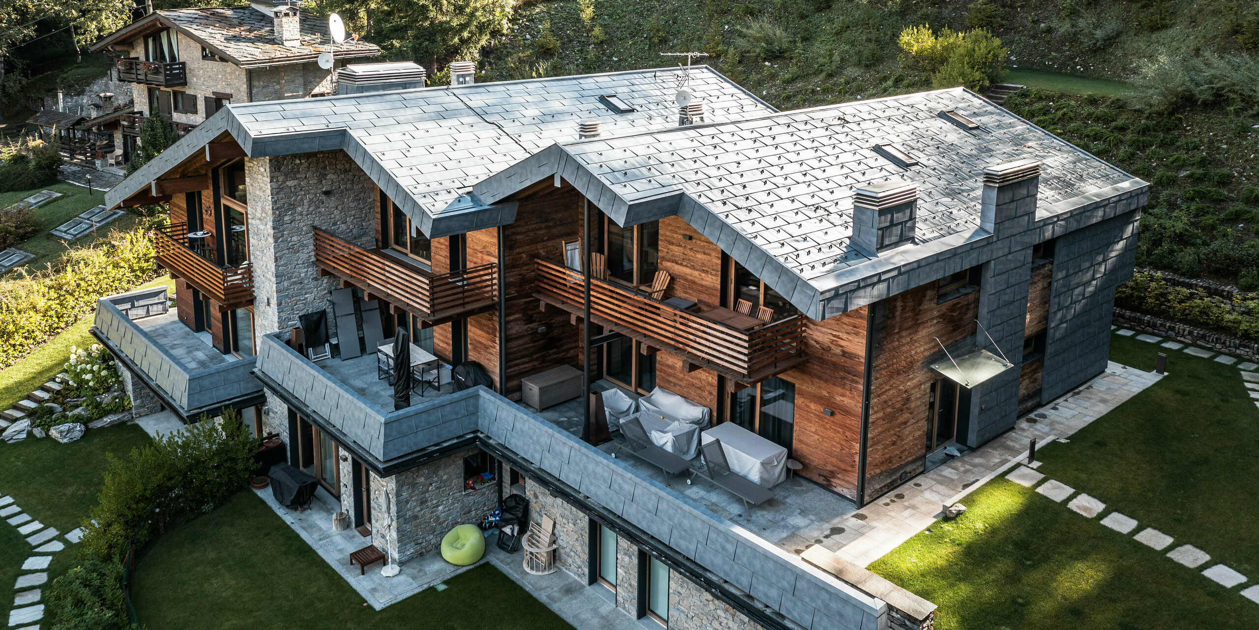 Luftaufnahme einer großzügigen Villa in Courmayeur, eingebettet in eine natürliche Waldlandschaft. Die Architektur kombiniert Holz, Stein und moderne PREFA FX.12 Aluminiumplatten in P.10 Steingrau. Die charakteristischen Balkone und Terrassen bieten weitläufige Außenbereiche zum Entspannen. Die Gartengestaltung mit präzisen Grünflächen und Steinelementen harmoniert mit der rustikalen und modernen Struktur der Villa und bietet einen atemberaubenden Blick auf die Alpen.