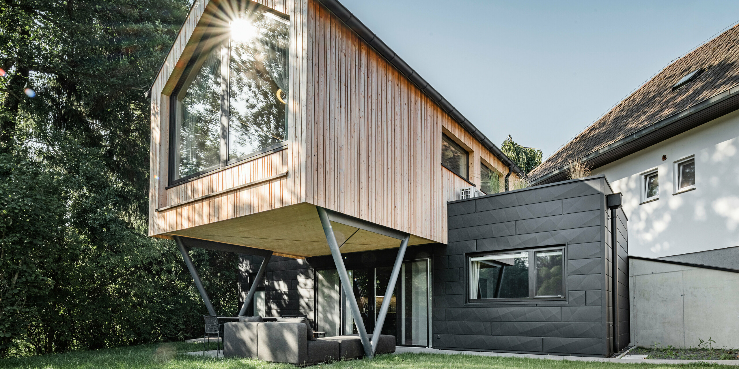 Das 'Woodstock Chalet 5' fügt sich mit seiner schwebenden Holzkonstruktion und den darunter befindlichen PREFA Siding.X Fassadenelementen in Anthrazit harmonisch in die natürliche Umgebung ein. Die großen Fenster des Obergeschosses reflektieren die umstehenden Bäume, die Sonne und den blauen Himmel. Die Fassadenelemente im unteren Bereich bieten einen modernen, industriellen Kontrast zur warmen Holzverkleidung oben und unterstützen das natürliche Design des Gebäudes. Die Sonne scheint durch die Blätter, was die Verbindung zwischen Architektur und Natur betont.