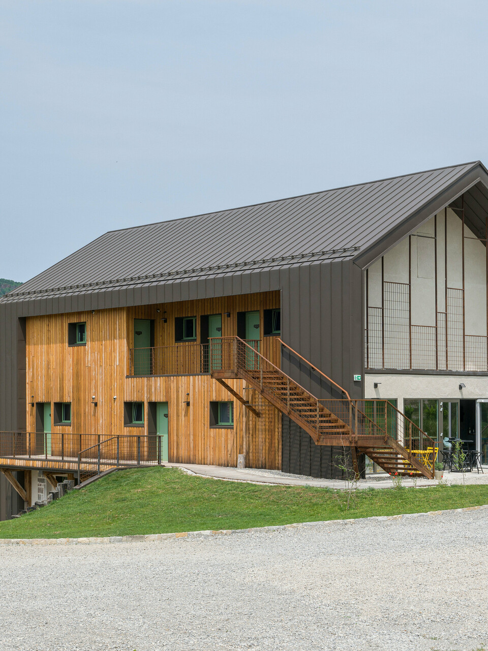 Eckansicht eines zeitgenössischen zweigeschossigen Hotels mit einer Hülle aus PREFA Aluminium und naturbelassener Holzfassade. Das Dach sowie Teile der Fassade wurden mit PREFALZ in P.10 Braun verkleidet. Hervorstechende Merkmale des Gebäudes sind große Fensterflächen sowie eine außenliegende Treppe, die zum oberen Stockwerk führt. Zudem verfügt das Hôtel en Tilleul über eine Terrasse im Erdgeschoss. Das Gebäude steht in einer idyllischen ländlichen Umgebung mit Blick auf sanfte Hügel. Am unteren Bildrand ist die Zufahrtsstraße aus Kies zu sehen. 