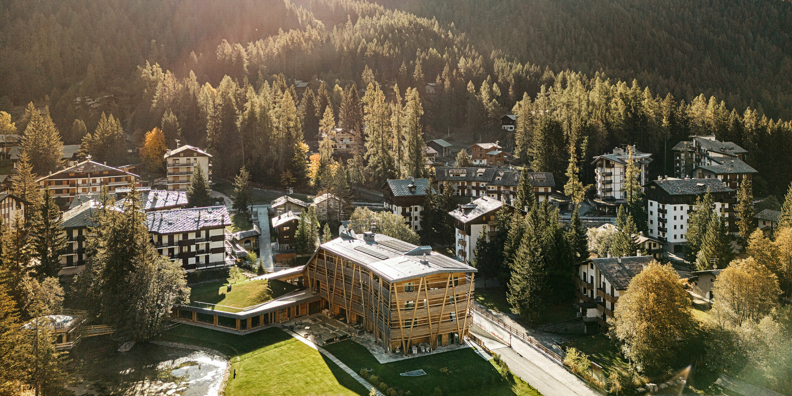 Luftaufnahme des Öko-Hotels 'Au Charmant Petit Lac' in Champoluc, eingebettet in die malerische Berglandschaft der italienischen Alpen. Das Hotel sticht mit seiner modernen Holzbauweise hervor, die sich harmonisch in das traditionelle alpine Dorfbild einfügt. Die umliegenden dichten Wälder und die ländliche Architektur der Region betonen die naturnahe Lage des Hotels, dessen Dach mit PREFALZ in P.10 Steingrau eine nachhaltige und ästhetische Bedachungslösung aufweist.