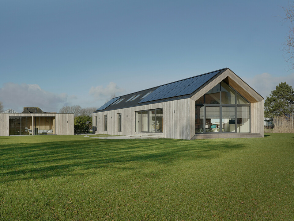 Weitwinkelaufnahme eines modernen Einfamilienhauses in Uithoorn, Niederlande, mit PREFALZ Winkelstehfalz-Eindeckung in Schwarzgrau und integrierter Photovoltaik-Anlage. Das Bild zeigt das gesamte Anwesen mit großzügigem Garten und einer Kombination aus großen Glasfronten und natürlichen Holzelementen an der Fassade. Die klare Linienführung und die hochwertigen, wetterbeständigen Materialien betonen die Nachhaltigkeit und das moderne Design des Hauses. Der einstöckige Bungalow wurde für die Wohnbedürfnisse einer Großfamilie erbaut.