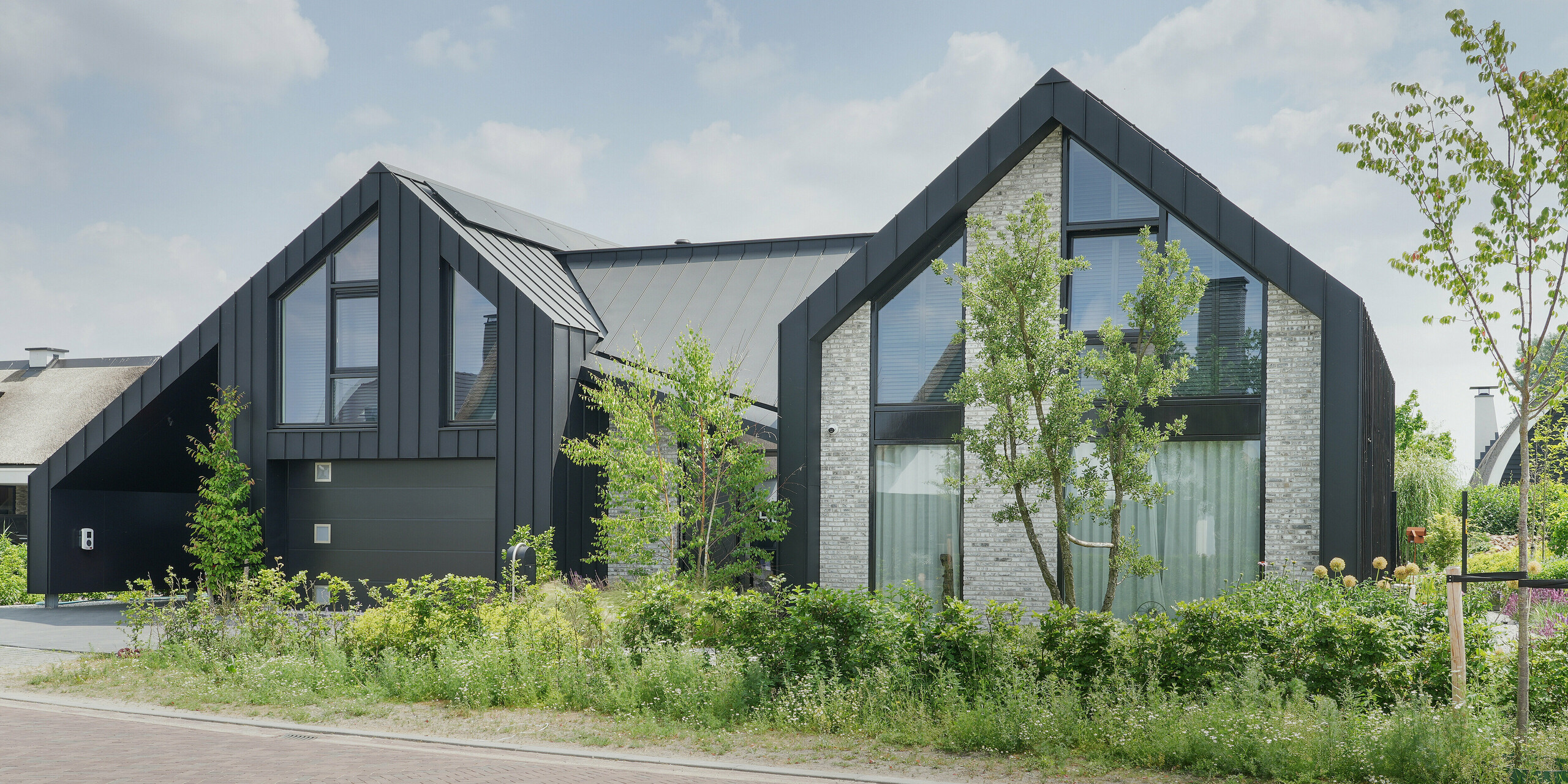 Ein Blick auf ein modernes Einfamilienhaus in Veenendaal, Niederlande, mit einem auffälligen schwarzen Dach und einer PREFALZ Fassade. Das Haus zeigt ein dynamisches Zusammenspiel von zwei Giebelstrukturen, die durch ein Flachdachelement verbunden sind. Akzente aus hellgrauem Naturstein an der Fassade lockern das dunkle Farbschema auf und sorgen für Struktur, die im Kontrast zu den großen Fensterfronten steht. Der Entwurf kombiniert klare, moderne Linien mit natürlichen Elementen, um eine Verbindung zwischen dem Wohnraum und der umgebenden Landschaft zu schaffen.