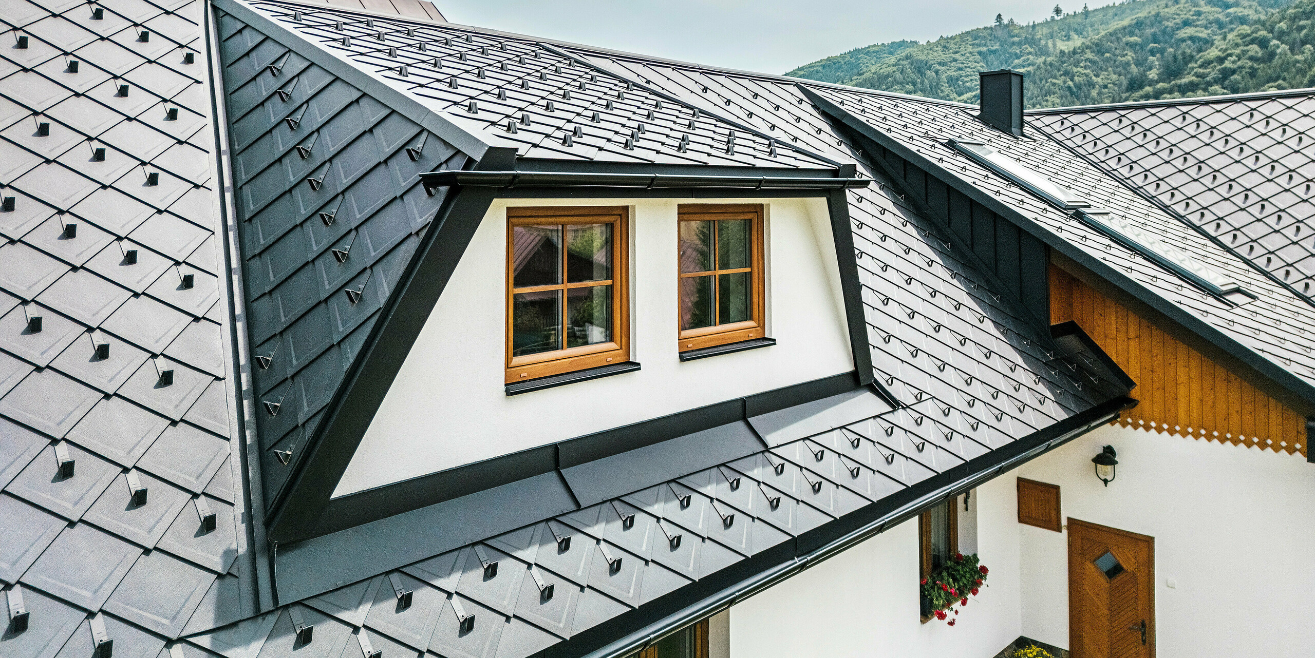 Nahaufnahme des Daches eines traditionellen Einfamilienhauses in Stara Bystrica, Slowakei, mit einer Trapezgaube, die mit PREFA Dachrauten 29 × 29 in P.10 Schwarz eingedeckt ist. Die langlebige Aluminiumdeckung in Schuppenoptik verleiht dem Dach ein modernes und elegantes Erscheinungsbild. Die perfekt integrierte Gaube und die Fenster mit Holzverkleidung fügen sich harmonisch in die Dachlandschaft ein und setzen architektonische Akzente.