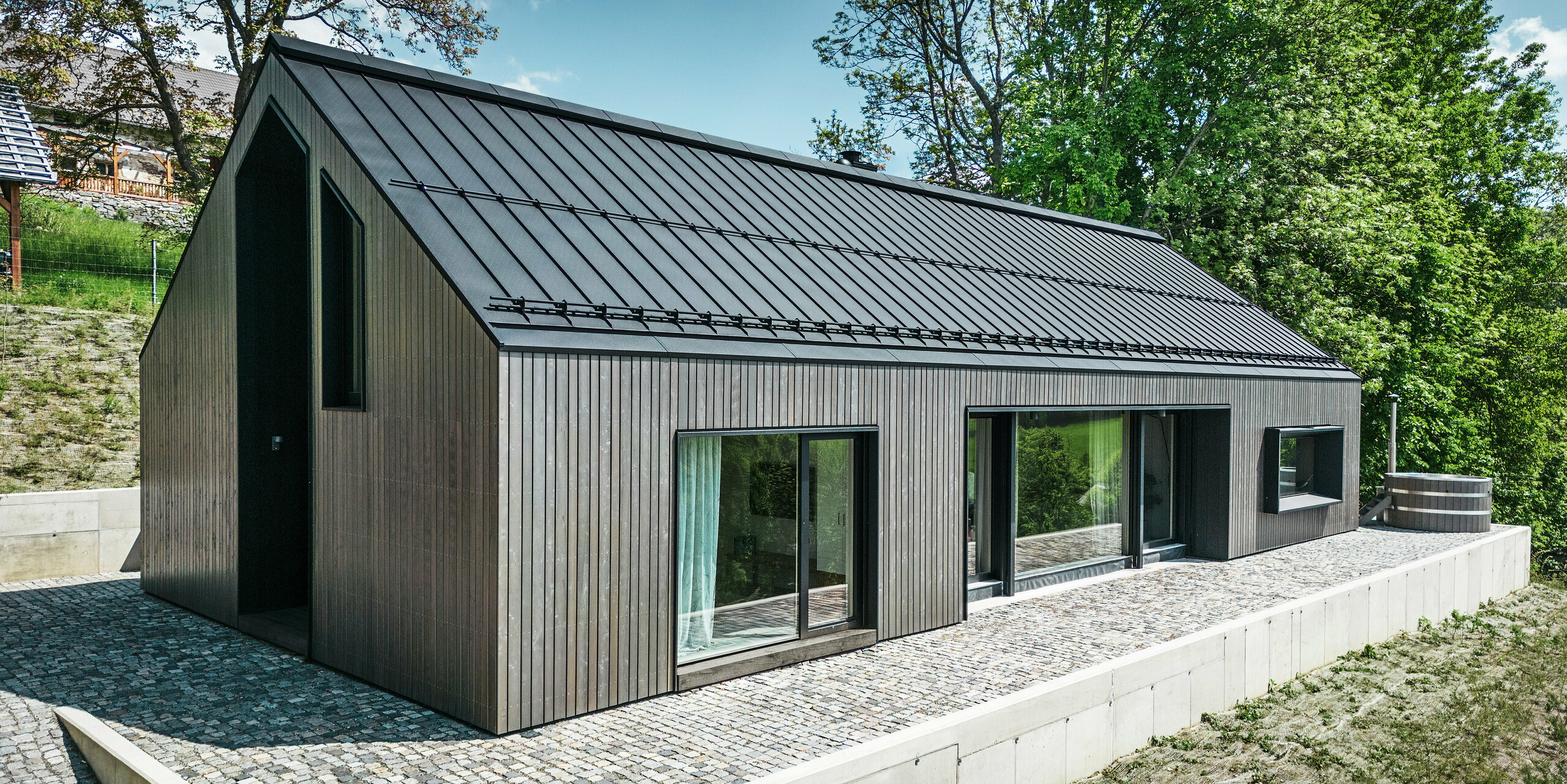 Vorderseite eines Holzmassivhauses in Kuncice, CZ. Das langlebige Aluminiumdach besteht aus PREFALZ in elegantem Schwarz. Das Haus verfügt über großzügige Fensterflächen und einen Whirlpool im Außenbereich.