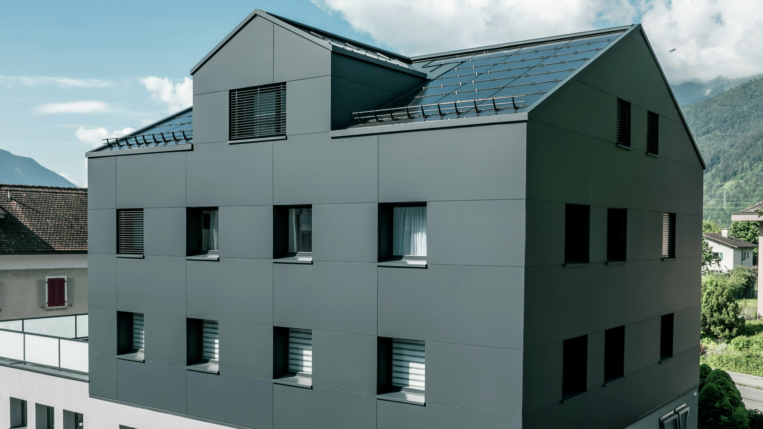 Modernisiertes Wohn- und Geschäftsgebäude nach der Fassadensanierung mit PREFABOND Aluminium Verbundplatten in Anthrazit. Die klare, elegante Linienführung der neuen Fassade verleiht dem Gebäude ein modernes Erscheinungsbild. Im Erdgeschoss befindet sich das Architekturbüro Voser. Die Umgebung mit Bäumen und Bergen rundet die harmonische Optik ab.